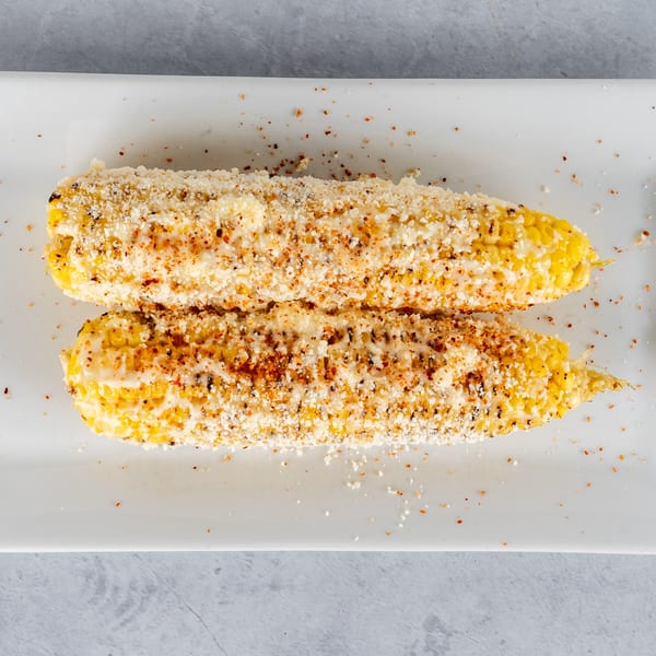 Two elotes, Mexican street corn, on a white plate, topped with cheese and spices.