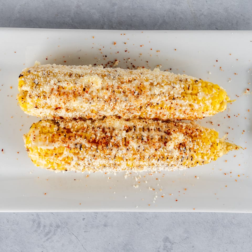 Two elotes, Mexican street corn, on a white plate, topped with cheese and spices.