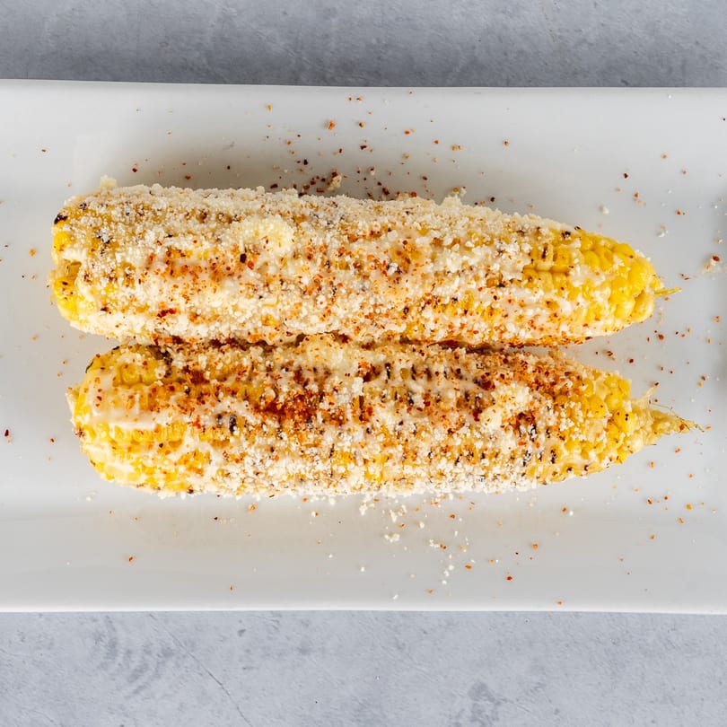 Two elotes, Mexican street corn, on a white plate, topped with cheese and spices.