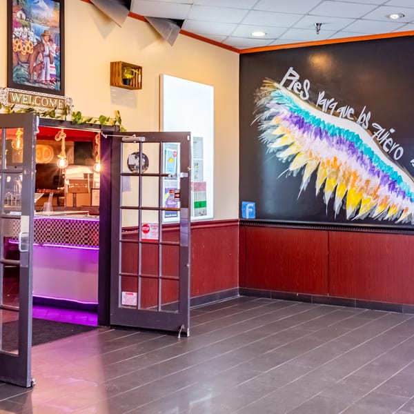 Restaurant entrance with a colorful wing mural, a welcome sign, and a bar with purple lighting.