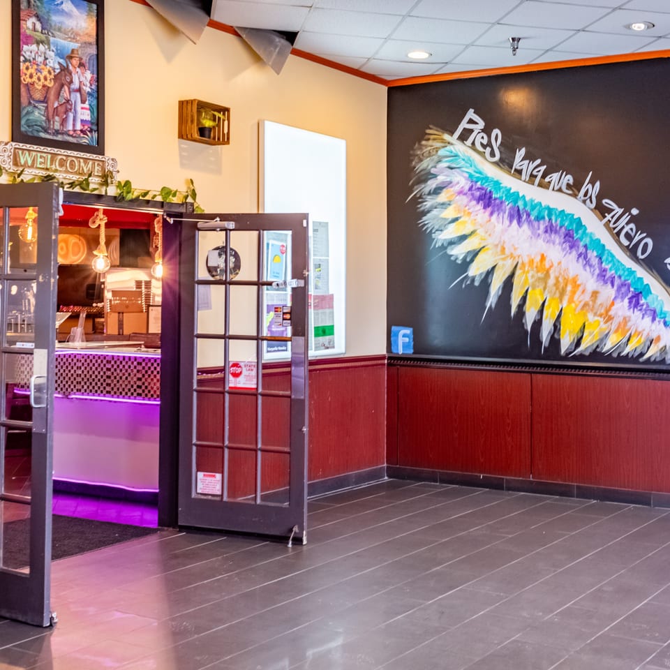 Restaurant entrance with a colorful wing mural, a welcome sign, and a bar with purple lighting.